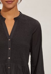 Svart knäppskjorta med en texturerad, smockad byst, volangkrage och långa ärmar; kontrasterande slät midja med en rynkad kjol.