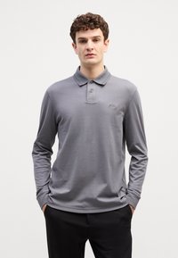 Polo majica s dugim rukavima od sive tkanine. Ima klasičan ovratnik, placket s tri gumba i mali logo na prsima. Glatka tekstura.