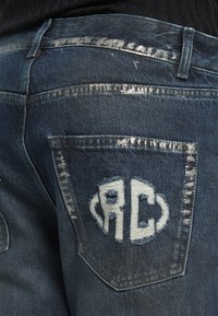 Denim jeans met een donkerblauwe wassing, met versleten patches en een wit geborduurd "RC" logo op de achterzak. Zilveren stiksels accentueren.