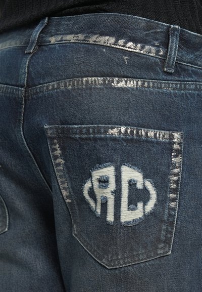 Denim jeans with a dark blue wash, featuring distressed patches and a white embroidered "RC" logo on the back pocket. Silver stitching accents.
