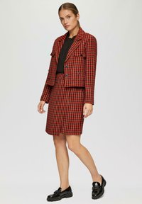 Rode en zwarte houndstooth-gepatroneerde blazer en rokset, met een cropped jack met dubbelgeknoopte sluiting en platte schoenen.