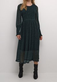 Mörkgrön långärmad klänning med smockad bodice och textil, genomskinlig tyg. Åtsittande ärmar med volangdetaljer vid ärmsluten. Bärs med svarta ankelstövlar.