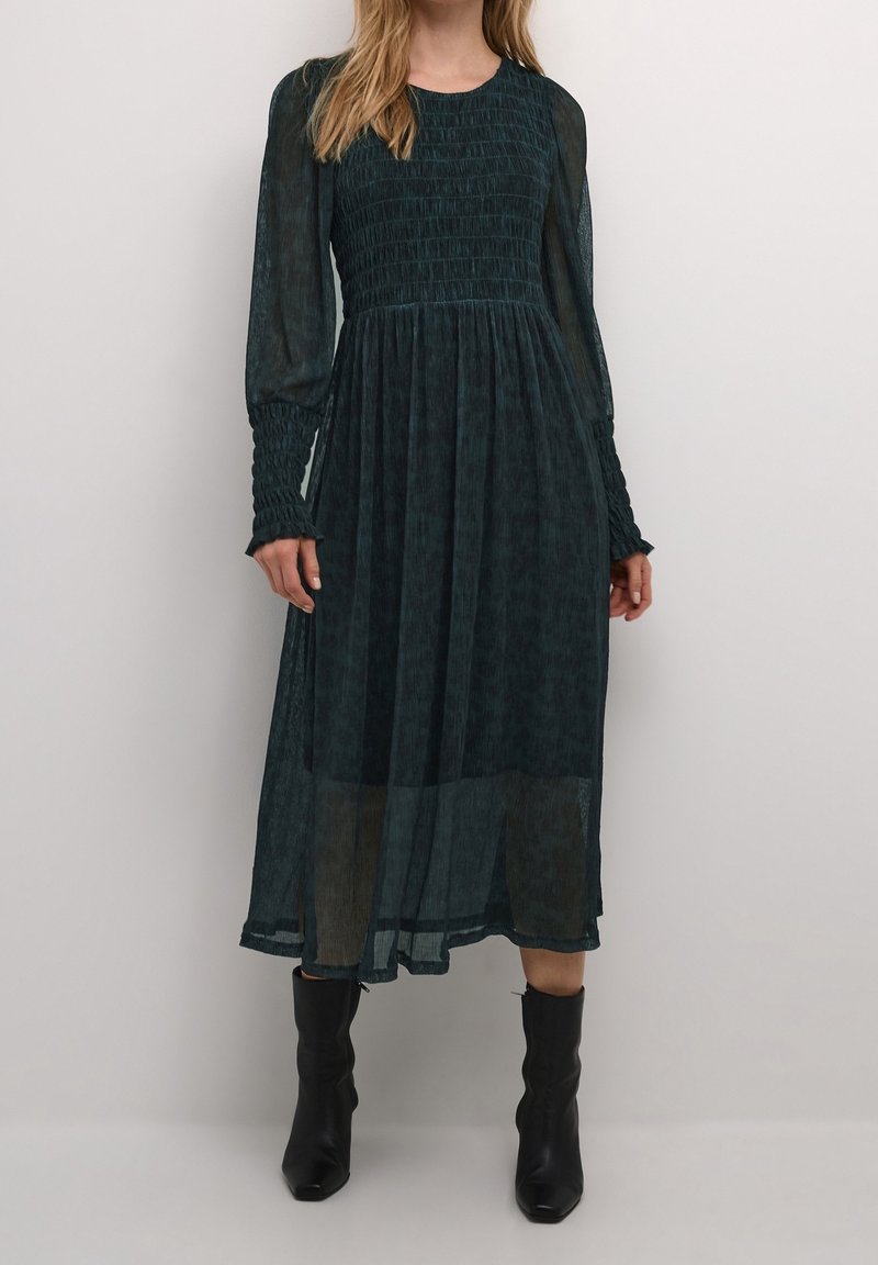 Mörkgrön långärmad klänning med smockad bodice och textil, genomskinlig tyg. Åtsittande ärmar med volangdetaljer vid ärmsluten. Bärs med svarta ankelstövlar.