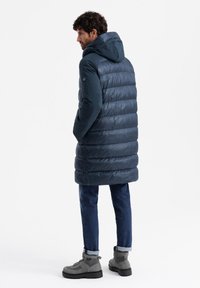 Marineblauer Puffermantel mit Kapuze, kombiniert mit einem gesteppten Design, Fronttaschen und langen Ärmeln. Trägt er über blauen Jeans und grauen Stiefeln.