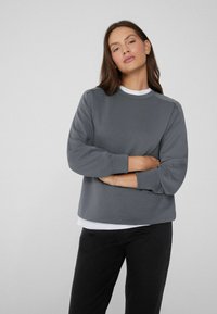 Grauer Pullover mit rundem Halsausschnitt, langen Ärmeln und lässiger Passform, über einem weißen Hemd getragen. Glatte Textur, minimales Design.