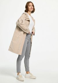 Beige waterdichte jas met een capuchon, knopen aan de voorkant en ritszakken. Draagt boven een witte top en grijze jeans, afgemaakt met beige sneakers.