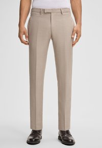 Beige maßgeschneiderte Hosen aus strukturiertem Stoff mit flachem Vorderteil, seitlichen Taschen und Gürtelschlaufen, kombiniert mit dunklen Schuhen.