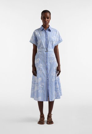 Vrouw draagt een midi-jurk met blauwe en witte strepen en witte bloemmotieven, korte mouwen, knopen aan de voorkant, een ceintuur in de taille en bruine sandalen met bandjes.