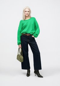 Πράσινο πλεκτό πουλόβερ, σκούρο denim φαρδύ παντελόνι, μαύρες γόβες με αιχμή μέχρι τον αστράγαλο και πράσινη τσάντα χειρός με διακοσμητικά στοιχεία.