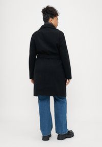 Zwarte wollen jas met een ceintuur in de taille, een notch-kraag en een getailleerde pasvorm, gedragen over blauwe wijde jeans en stevige zwarte schoenen.