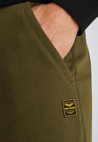 Olijfgroene sweatpants met een gestructureerde afwerking, voorzien van een zijzak en een geel en zwarte merklabel op de dij.