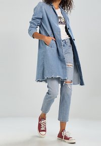 Abrigo largo de denim en azul claro, con un bajo deshilachado, dos bolsillos laterales, combinado con una camiseta gráfica blanca y jeans rotos de azul claro, terminado con zapatillas burdeos.