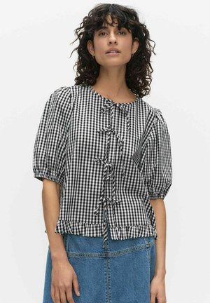 Vrouw met krullend donker haar draagt een zwart-wit geruite blouse met pofmouwen, voorstrikjes en een gerimpelde zoom, gecombineerd met een blauwe spijkerrok.