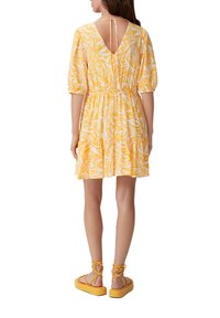 Gelbes, kurzärmliges Kleid mit floralem Muster, V-Ausschnitt hinten, tailliert und mit geschichtetem Rock. Kombiniert mit gelben Sandalen mit Riemen.