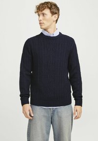 Navy blue cable-knit sweater with ribbed cuffs and hem, round neckline, and textured pattern. Worn over a light blue collared shirt.