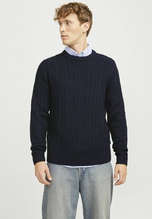 Navy blue cable-knit sweater with ribbed cuffs and hem, round neckline, and textured pattern. Worn over a light blue collared shirt.