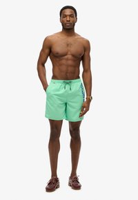 Costume da bagno verde con vita regolabile, con una stampa del logo blu sul lato. Il modello ha una corporatura snella e è a piedi nudi.