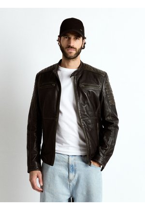 Homme portant une veste en cuir noire, un t-shirt blanc, un jean bleu clair et une casquette noire, debout devant un fond blanc uni.