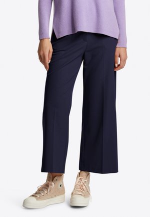 Femme portant un pantalon large cropped bleu marine, un pull violet clair, et des baskets montantes beige avec des semelles et des lacets blancs.