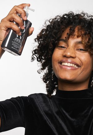 Haut en velours noir à manches courtes, tenant un flacon vaporisateur transparent avec un embout incurvé. L'étiquette affiche le nom du produit et les instructions.