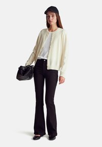 Kremowy kardigan z guzikami, biała koszula, czarne rozkloszowane dżinsy, czarne baleriny i czapka w kolorze czarnym. Niesie torebkę z teksturowanej czerni.