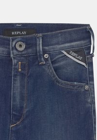 Jeans de ganga azul escuro com um bolso frontal com fecho de botão e costura tonal. A etiqueta diz "REPLAY." Textura suave.