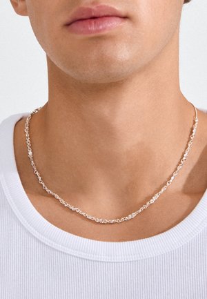 Primo piano di collo e parte inferiore del viso con una collana a catena d'argento intrecciata e una maglietta bianca a coste con scollo tondo.