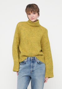 Žlutohnědý pletený svetr s rolákem a oversized rukávy, měkká textura a žebrovaný lem, spárovaný se světle modrými džíny.