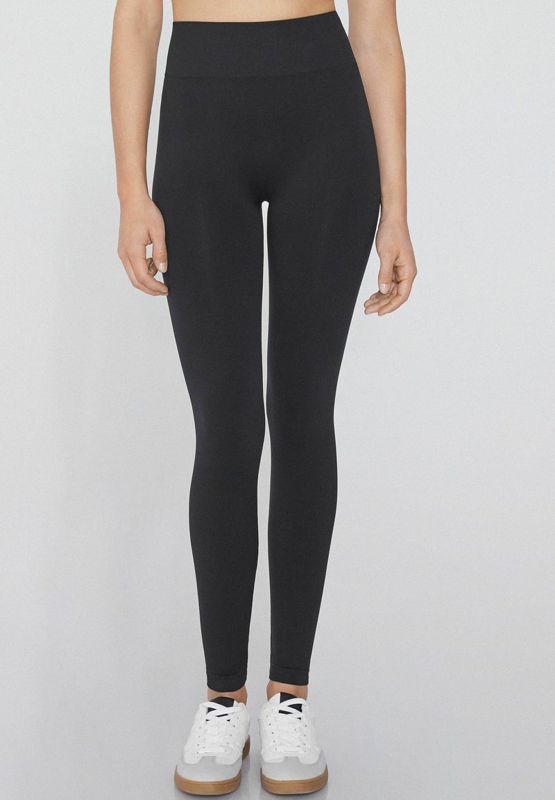 Schwarze Leggings mit einem hohen Bund, aus dehnbarem Stoff, mit einer glatten Textur und einer enganliegenden Passform, kombiniert mit weißen Sneakern.