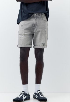 Persoană care poartă pantaloni scurți de denim gri deschis, răsuciți și rupți, adidași negri și albi, șosete albe și o cămașă închisă la culoare, cu brațele încrucișate.