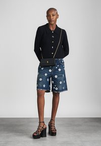 Vrouw draagt zwarte trui met knopen, denim shorts met witte bloemen, zwarte crossbodytas en zwarte platformsandalen, staand tegen een witte muur.