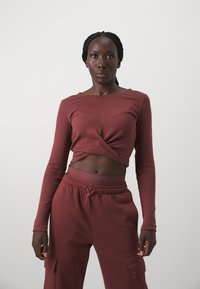 Burgundi hosszú ujjú felső, csavart elülső dizájnnal, hozzáillő joggernadrággal, oldalsó zsebekkel és elasztikus derékkal. Sima textúra.