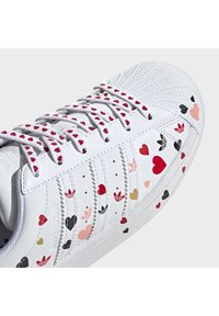 Sneakers en cuir blanc avec des motifs en forme de cœur en rouge, rose, noir et doré, dotés d'un système de laçage et d'une semelle en caoutchouc texturée.