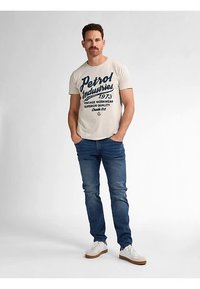 Beigenvärinen puuvillainen t-paita, jossa on tummansininen teksti ja logo, yhdistetty sinisiin farkkuihin ja valkoisiin lenkkareihin. Malli seisoo rennosti käsien ollessa taskuissa.