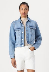 Chaqueta de mezclilla corta en azul claro con cierre de botones, dos bolsillos en el pecho y un cuello clásico. Lleva puesta una camiseta de manga larga en un tono neutro.