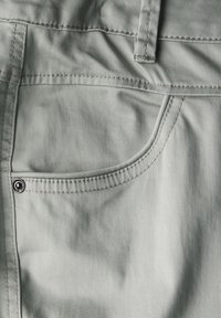 Jasnozielony materiał z bliska przedstawiający kieszeń spodni, z metalowym zapięciem na zatrzask, przeszyciem w górnej części oraz gładką teksturą z lekkim połyskiem.