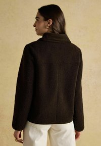 Hnedá textúrovaná fleecová bunda s vysokým golierom a voľným strihom, obsahujúca priamé rukávy a minimalistické dizajnové prvky.