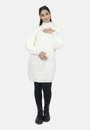 Kvinde i hvid strikket sweaterkjole og sorte leggings, stående med hænderne på brystet og maven, smilende blidt.