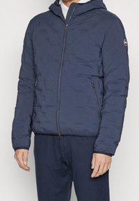 Man draagt een marineblauwe gewatteerde jas met capuchon en ritssluitingen, en een bijpassende marineblauwe broek, staand tegen een effen achtergrond.