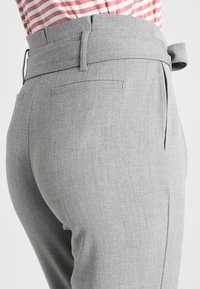 Grijze broek van een gestructureerde stof, met een omgevouwen tailleband, zijzakken en een riemdetail. Gestreepte shirt zichtbaar erboven.