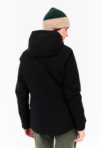 Vrouw in een zwarte hoodie, met een groen en beige gebreide beanie en olijfgroene broeken, gezien vanaf de achterkant tegen een witte achtergrond.