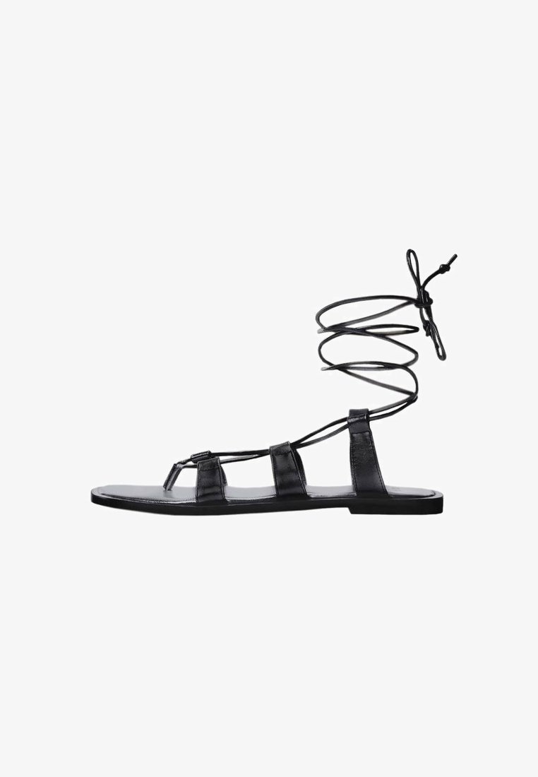 Sandales en cuir noir avec semelles plates, comprenant trois parties de lanières et un lien en spirale autour de la cheville. Texture lisse et design minimaliste.
