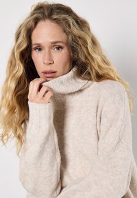 Jasnobeżowy sweter z golfem, dziany, z prążkowanymi mankietami, teksturowanym wzorem i luźnym krojem. Materiał wydaje się miękki i ciepły.