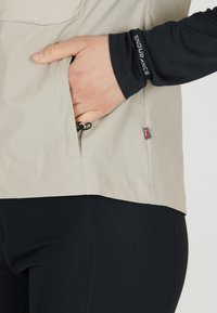 Leichte beige Jacke mit strukturiertem Finish, ausgestattet mit einer Seitentasche und einem verstellbaren Zugverschluss. Getragen über einem schwarzen Langarmoberteil.
