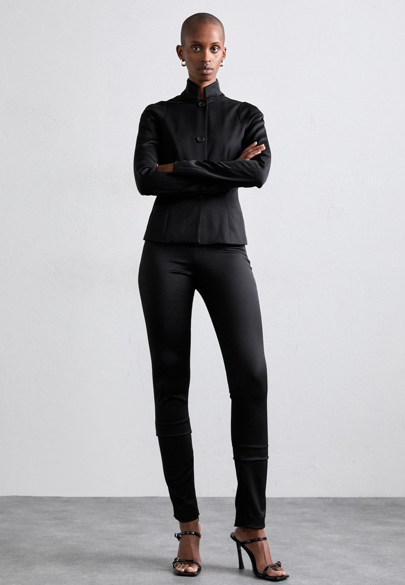 Black tailored jacket with a high collar and button closure, paired with fitted black trousers and strappy black high-heeled sandals.