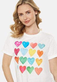 Hvid T-shirt med et gentaget mønster af akvarel hjerter i pink, blå, orange, gul, grøn og aqua. Korte ærmer.