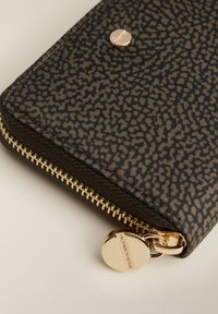 Angolo di un portafoglio texture marrone e nero con cerniera dorata e tirazip rotondo marchiato su una superficie chiara.