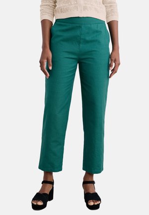 Femme portant un pantalon droit bleu sarcelle et des talons ouverts noirs, avec un pull en maille crème court sur un fond uni.