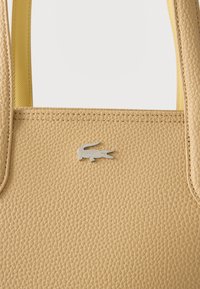 Μπεζ υφασμάτινη δερμάτινη τσάντα με διπλές λαβές και μικρό ασημένιο λογότυπο κροκοδείλου κεντραρισμένο στο μπροστινό μέρος.