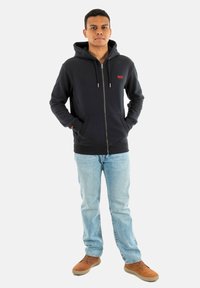 Marineblauwe zip-up hoodie met trekkoorden, voorzakken en een klein rood logo, gecombineerd met lichtblauwe jeans en bruine schoenen.
